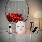 Facial mask with red light, remote control, and other devices on a surface with a blurred heart-shaped light in the background.